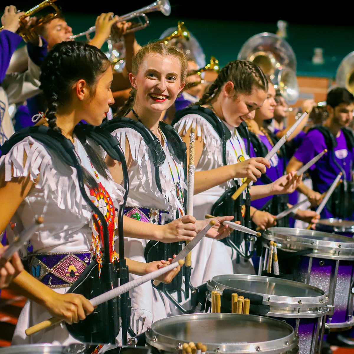 As they await their cue, varsity twirler Sophia Totten ('26) smiles at fellow twirler Paulina Torres ('25) as they set to perform with the drum line during the third quarter of the football game with Montgomery on Friday, Nov. 1, 2024. The line traditionally plays near the student section while the rest of the band returns to their seats and sections following halftime. Along with  Gracelynn Snider ('25), the three twirlers regularly trade out their batons for drums sticks and perform during the cadences. (Izzy Zeig/NDN Press)